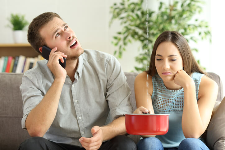 Concerned young couple at home, woman holding a red bowl under a leaking ceiling while man talks on a cell phone, seeking help froma plumber.