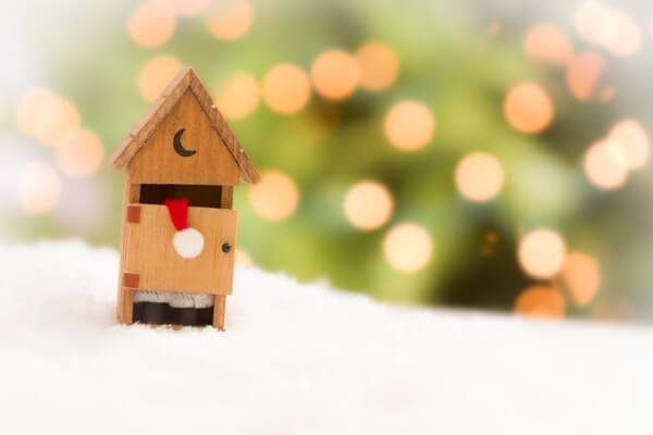 Miniature Santa figure inside a small wooden outhouse surrounded by snow with soft holiday lights in the background.